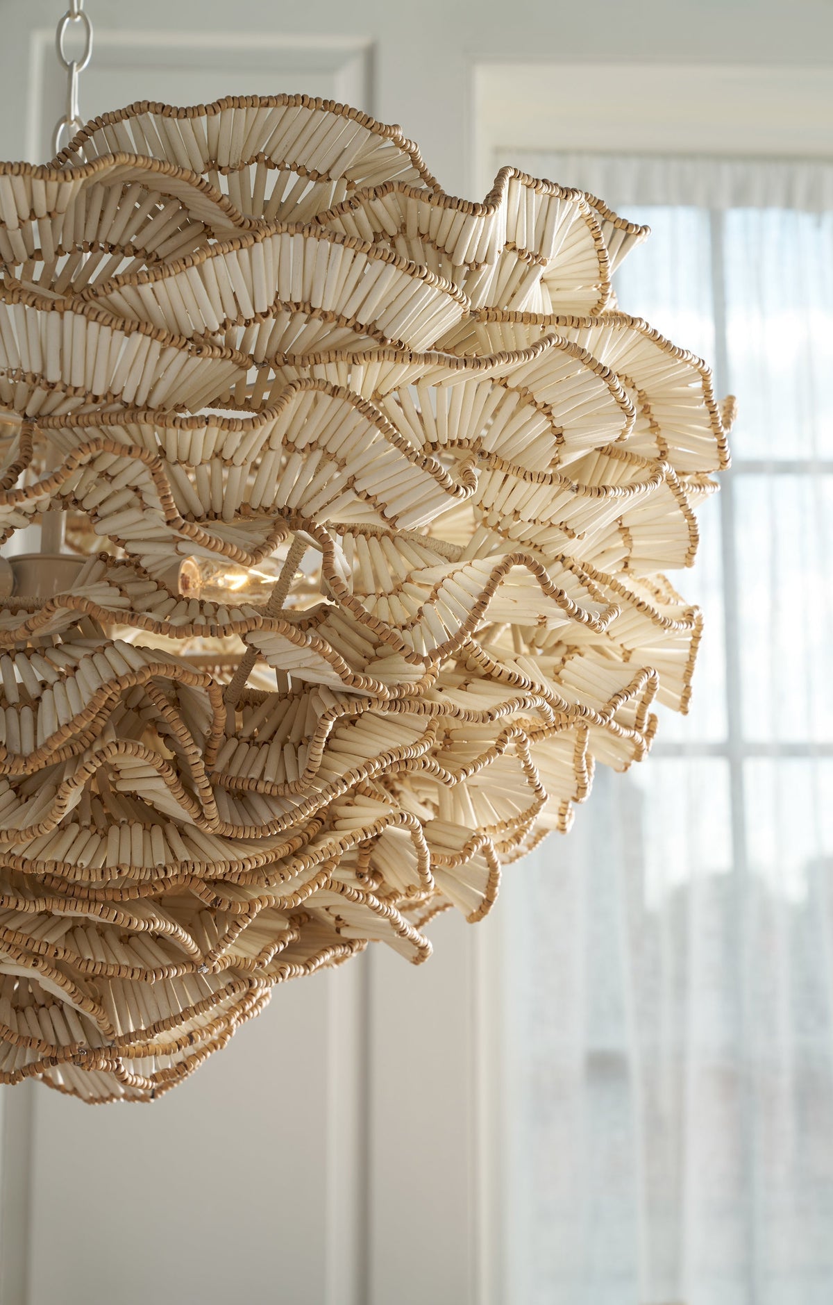 Close-up of a hand-beaded coconut shell chandelier with wavy, ruffled tiers, hanging near a sunlit window with sheer curtains.