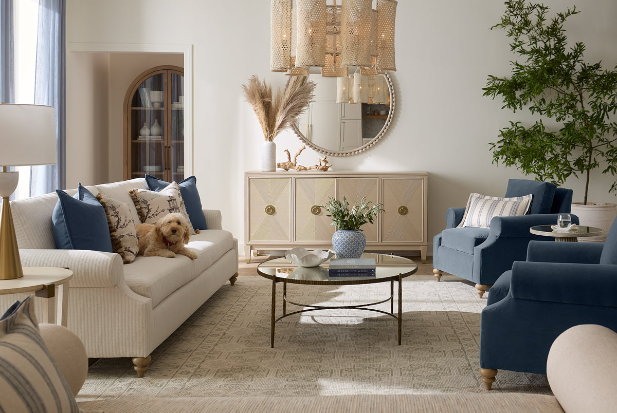 Gabby cream sofa and navy velvet lounge chairs in a bright coastal living room at Gabriella White, styled with a woven sideboard, brass coffee table, rattan chandelier, and layered blue and neutral accessories in a welcoming Coast-pillar interior.