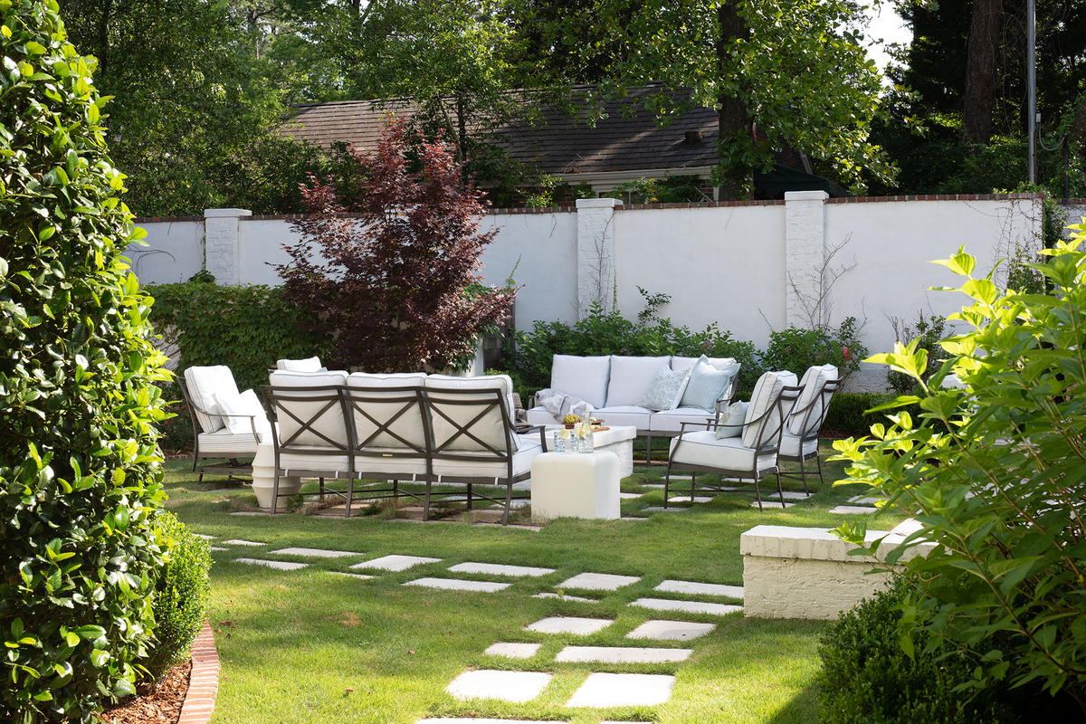 Summer Classics cast aluminum outdoor lounge chairs and sofa with crisp white cushions gathered around a stone coffee table in a walled garden, surrounded by boxwood, stepping stone pavers, and mature trees.